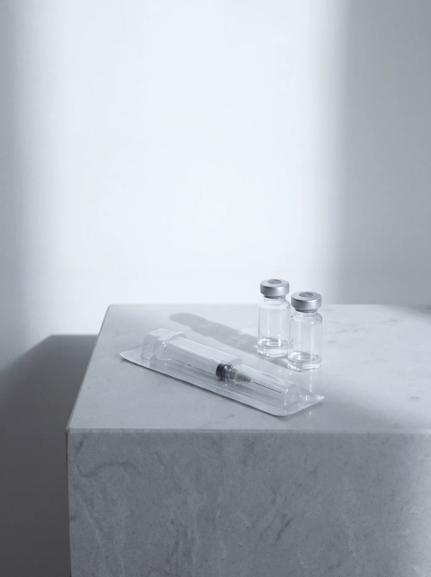 A syringe in its sealed tray beside two glass vials on stone