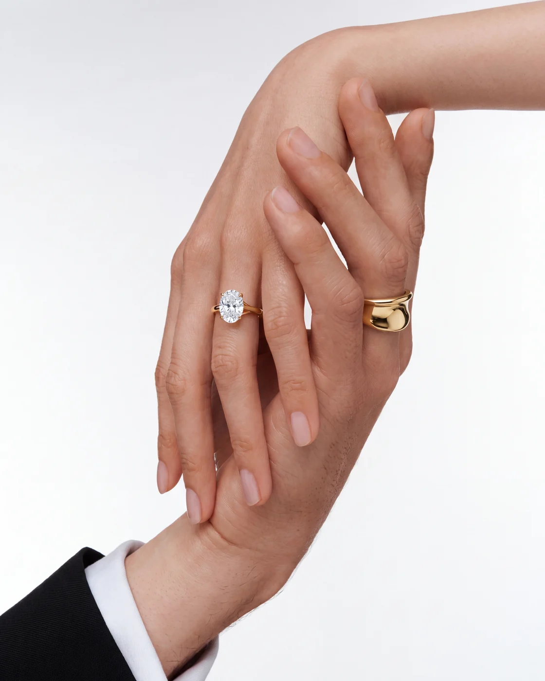A woman in a black blazer rests her hand on her cheek, wearing an oval diamond engagement ring and a wide gold band from the OVELLE jewellery collection