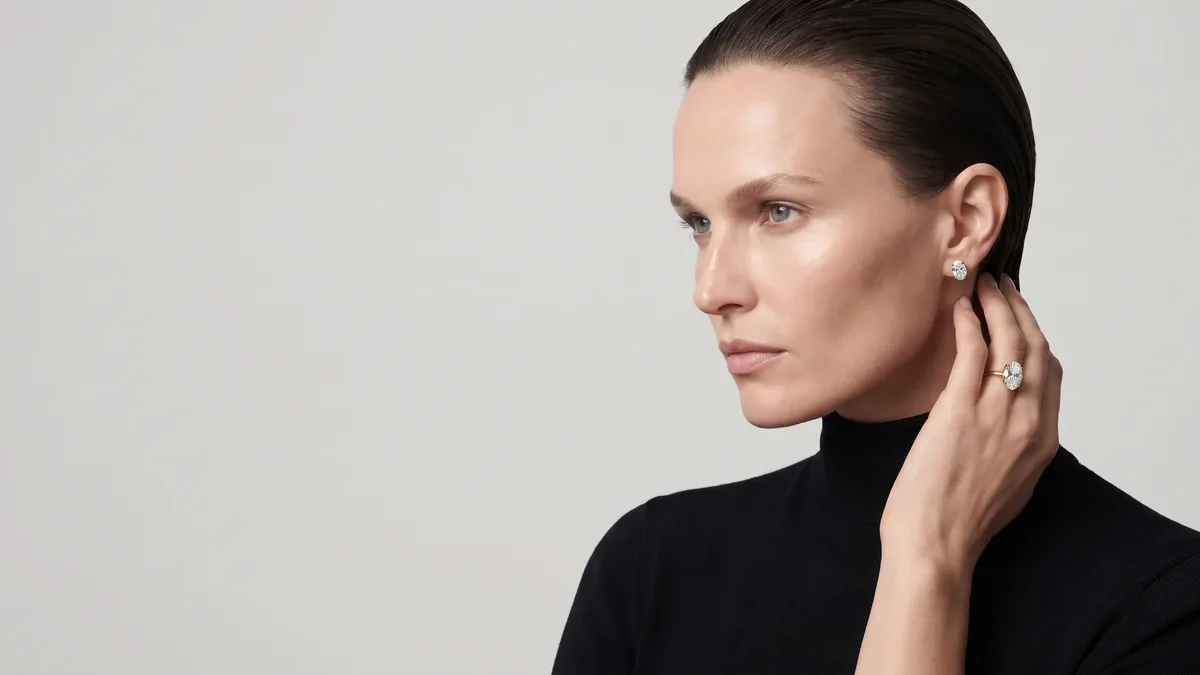Editorial image for OVELLE, a fictional jewellery house from the Libra Studio client-workspace concept: a model in a black turtleneck in profile, wearing an oval diamond stud and an oval diamond ring on the hand raised to her neck, against a light grey backdrop